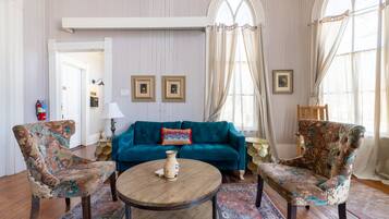 A room with a teal sofa, two patterned armchairs, a wooden coffee table, and a large window with curtains.