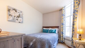 Une chambre à coucher avec un lit, une table de chevet, une lampe, une fenêtre avec des rideaux et un tableau au mur.