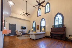 Un salon spacieux doté de planchers en bois franc, d’un grand canapé, d’un piano et de nombreuses fenêtres surmontées d’arcs.