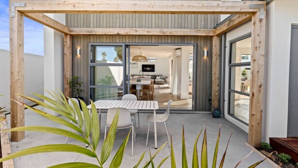 Una casa moderna con un patio amueblado con pérgola de madera, un comedor y una cocina visible a través de puertas de vidrio.