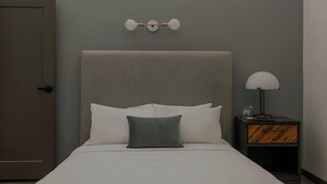 A neatly made bed with white and gray bedding, a gray headboard, and a bedside table with a lamp and a small decorative item.