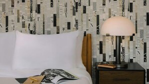 Un lit bien fait, avec un magazine et un stylo posés dessus, une lampe sur une table de chevet et un mur recouvert d’un papier peint à motifs de journaux.
