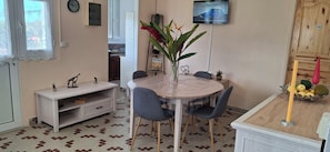 Ein Essbereich mit einem runden Tisch, Stühlen, einer Vase mit Blumen, einem Fernseher an der Wand und einer Küche im Hintergrund.