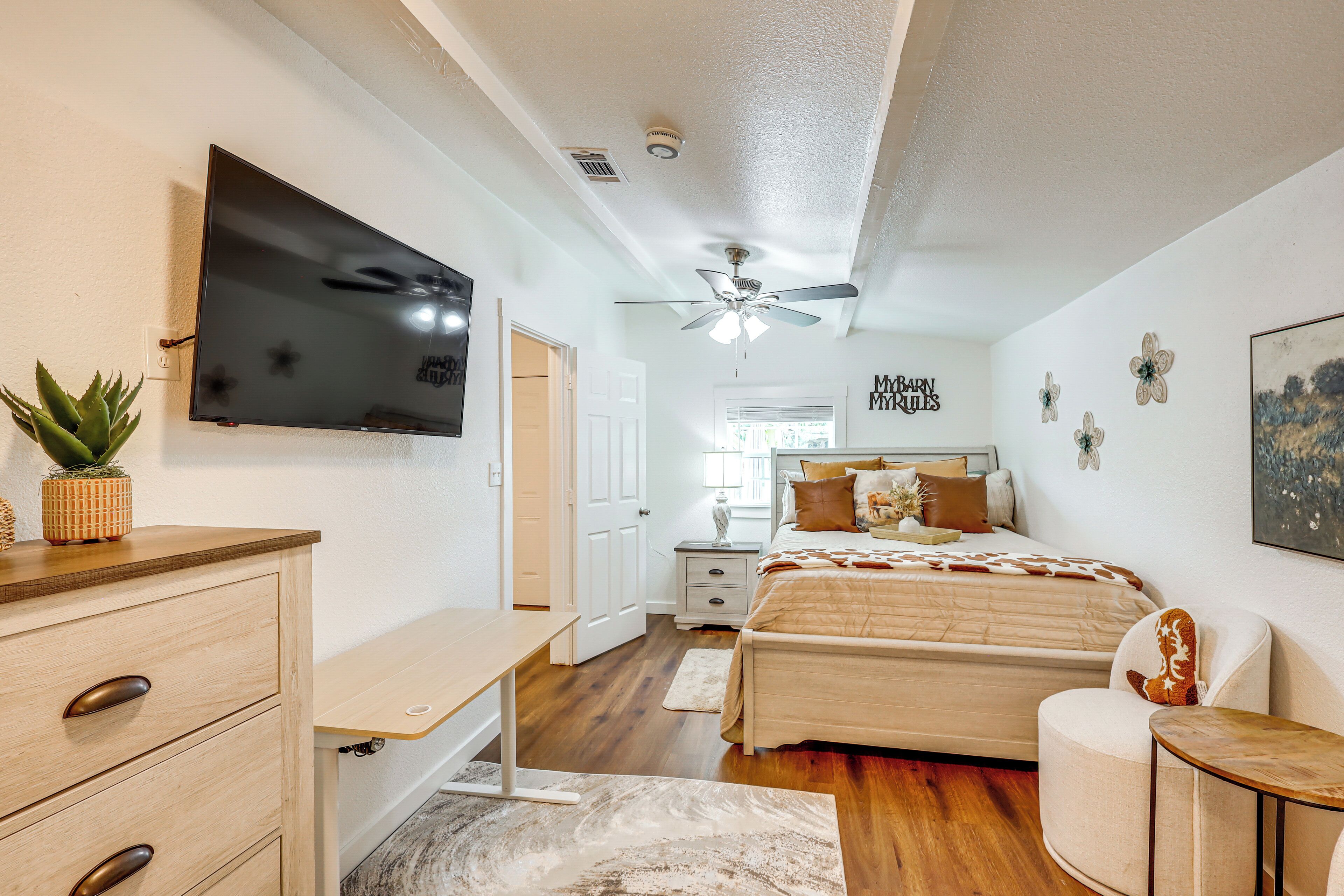 A bedroom with a bed, a desk, a chair, a TV, a ceiling fan, and a wall with a sign.