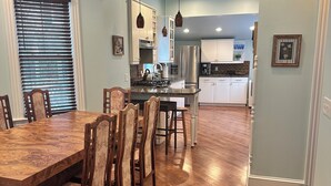 A spacious kitchen with a central island, wooden dining table, and chairs. There are pendant lights above the island and a refrigerator in the background.