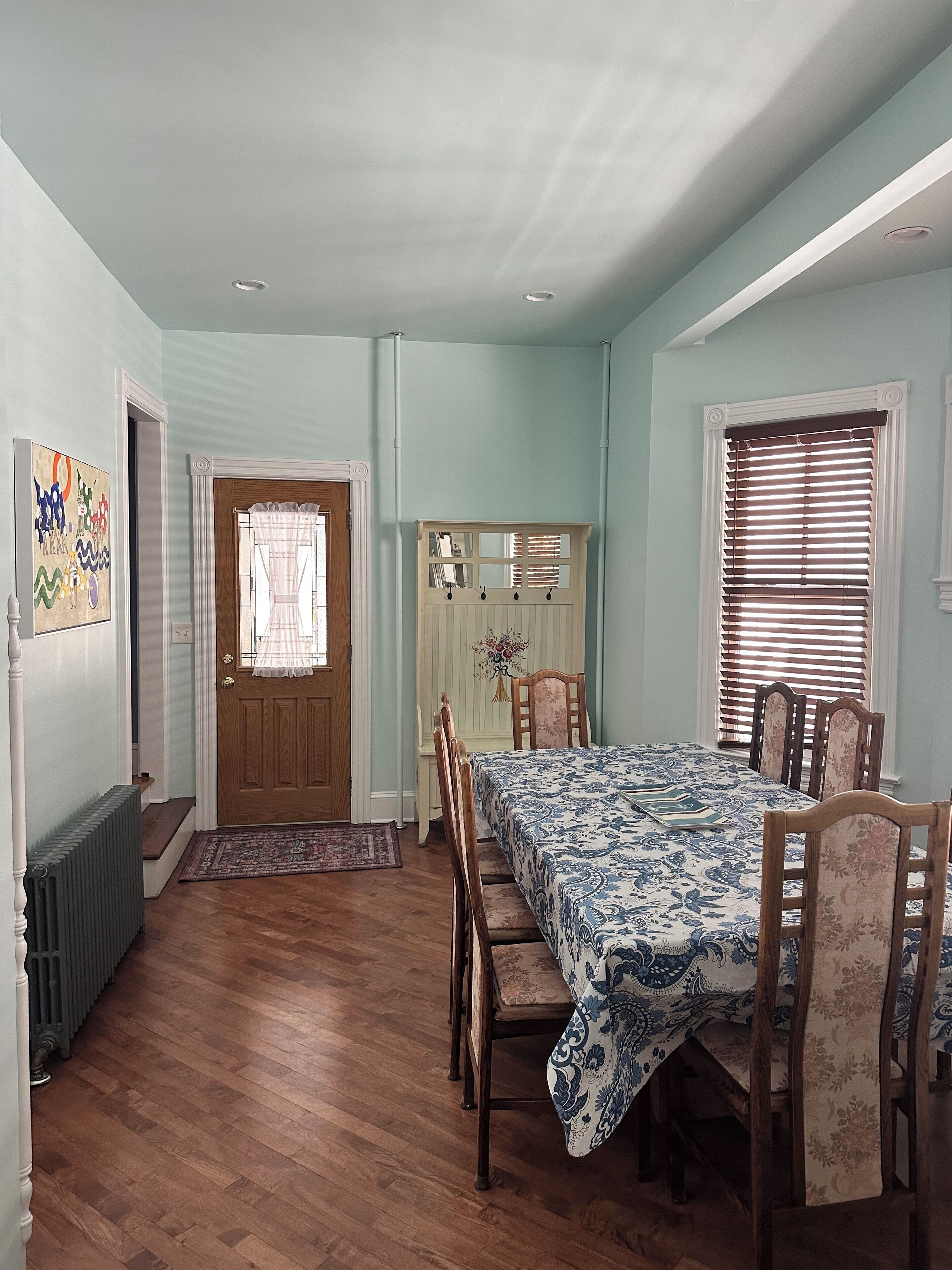 A dining room with a table set for four, flanked by wooden chairs. A door with a window and a coat rack with a mirror are visible.
