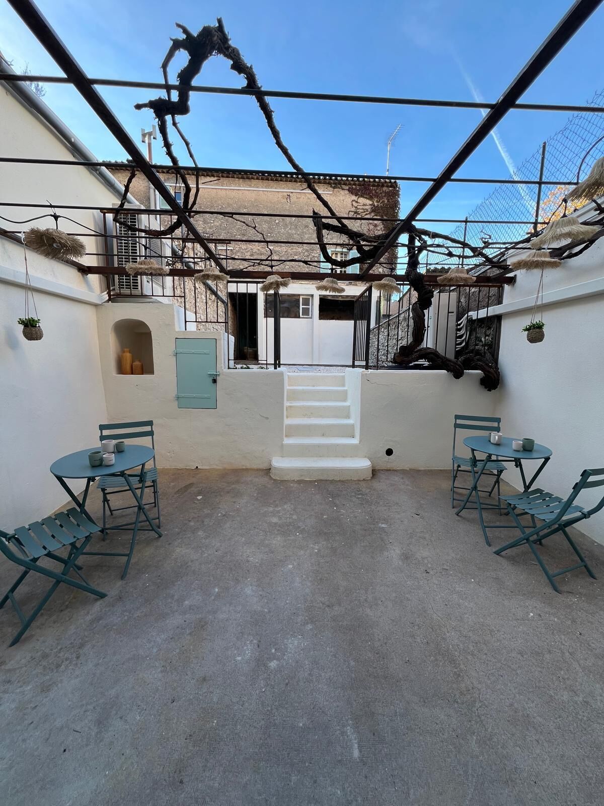 A courtyard with a white staircase leading to a door, surrounded by walls and a metal fence. There are two tables with chairs and hanging plants.