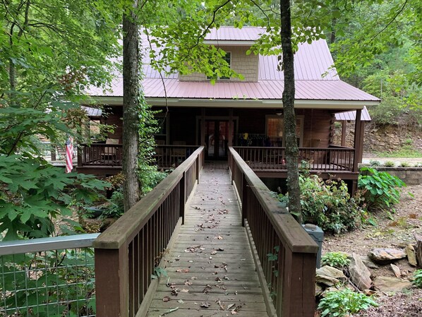 A wooden bridge leading to a cabin with a metal roof, surrounded by trees and a stone wall.