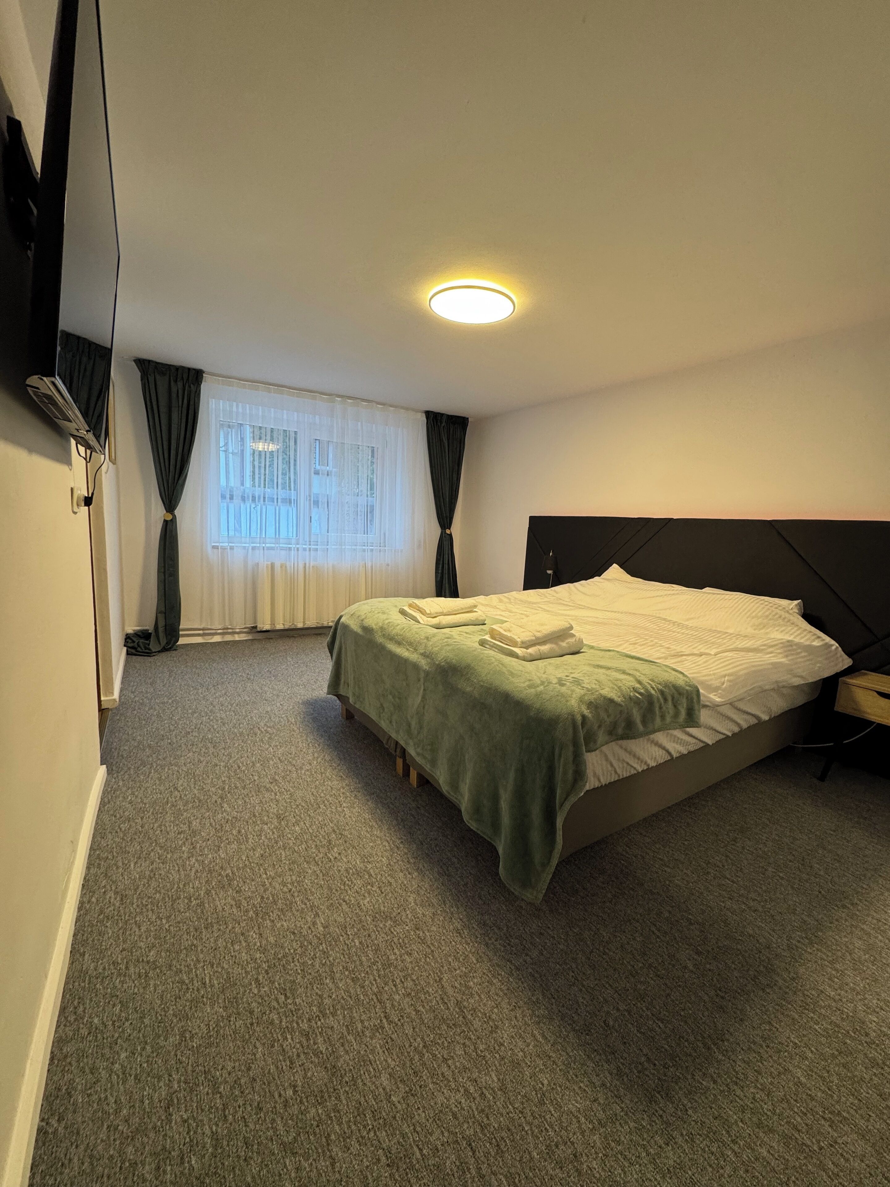 Ein Hotelzimmer mit Bett, Nachttisch, Lampe, einem Fenster mit Vorhängen und einer Deckenleuchte.