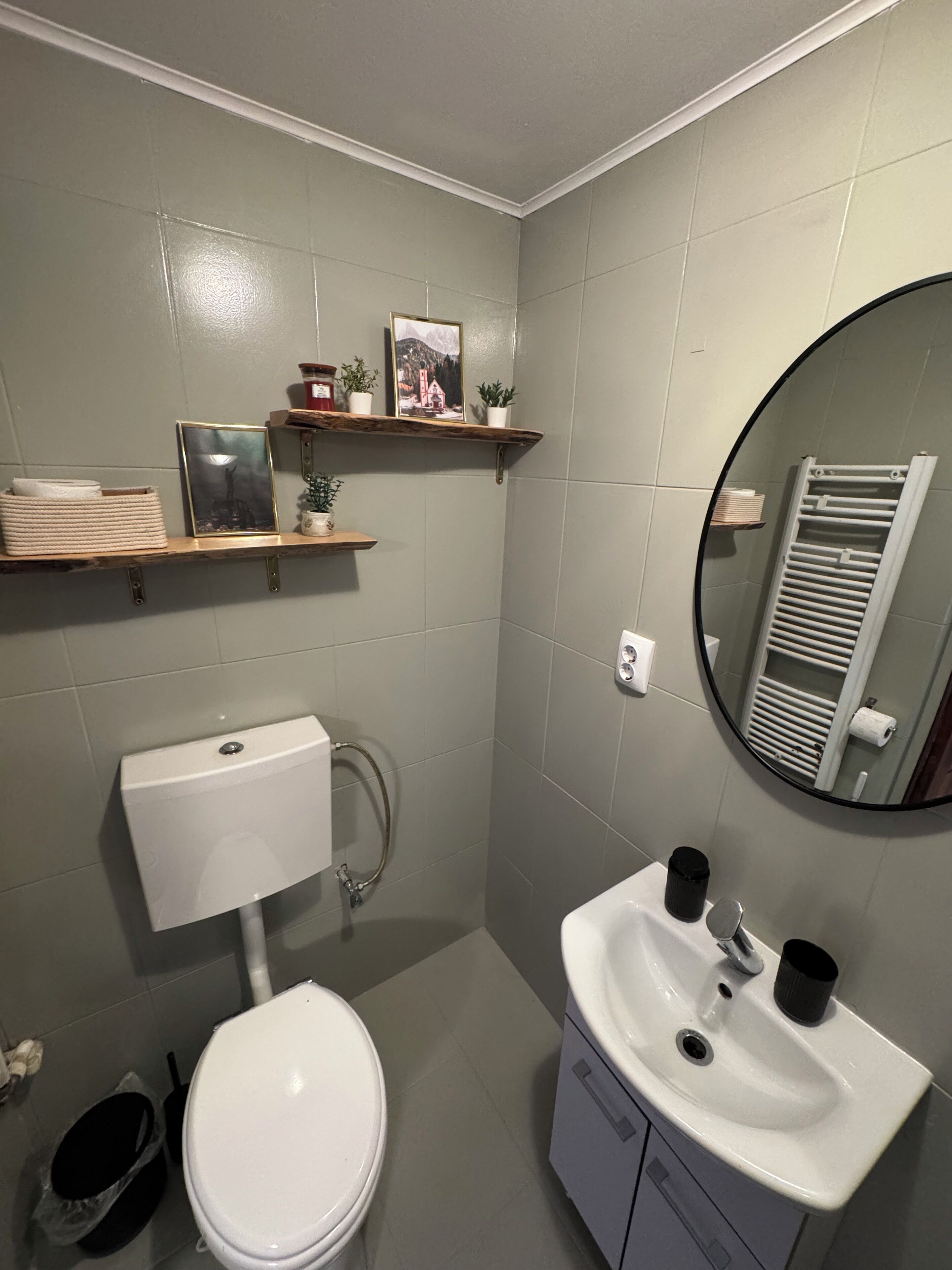 Casa de banho com sanita, lavatório e espelho. Prateleiras com objetos decorativos e um porta-toalhas.