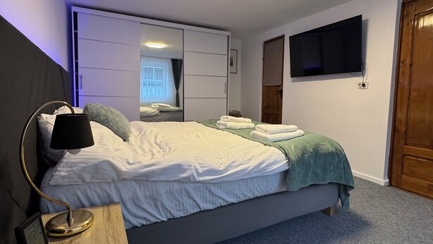 A bedroom with a bed, bedside table, lamp, wardrobe, and a television on the wall.
