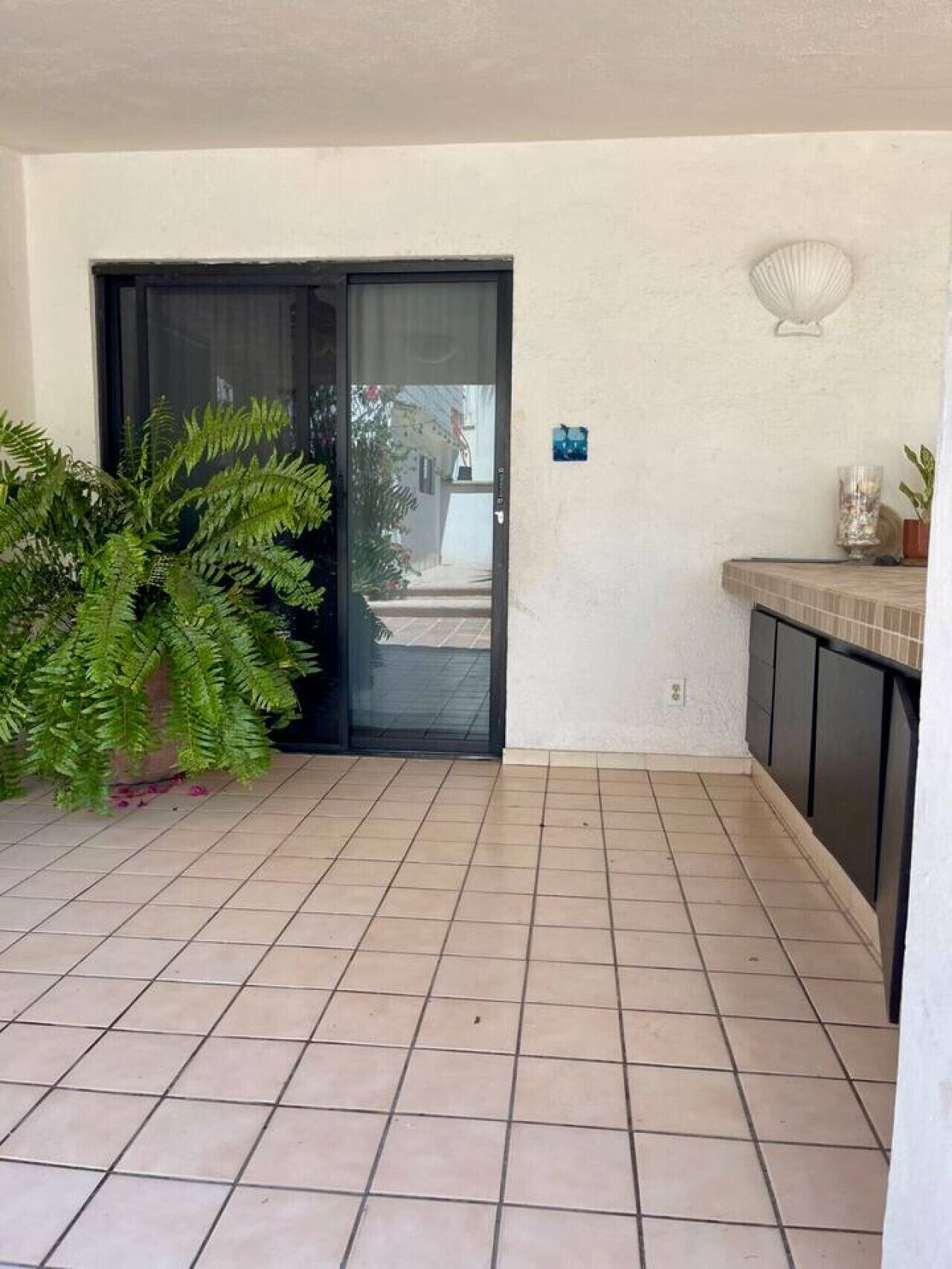 Ein gefliester Balkon mit einer Schiebeglastür, einem Topf mit einem Farn und einem kleinen, an der Wand befestigten Regal mit Dekorationen.