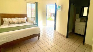 Quarto de hotel com cama, ventoinha no teto, janela com cortinas, banheiro com pia e espelho, e piso revestido de azulejos.