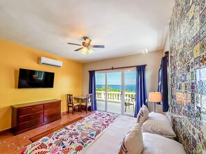 Ein Hotelzimmer mit Bett, Fernseher, Deckenventilator, Schreibtisch mit Stühlen, Meerblick durch große Fenster und einer farbenfrohen Wand.
