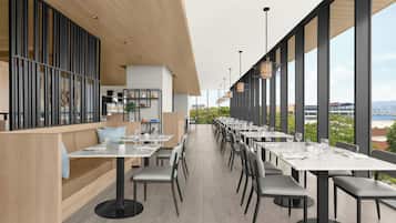 Moderni ravintola, jossa on pitkiä pöytiä ja tuoleja, suuret ikkunat ja näkymä kaupunkiin.