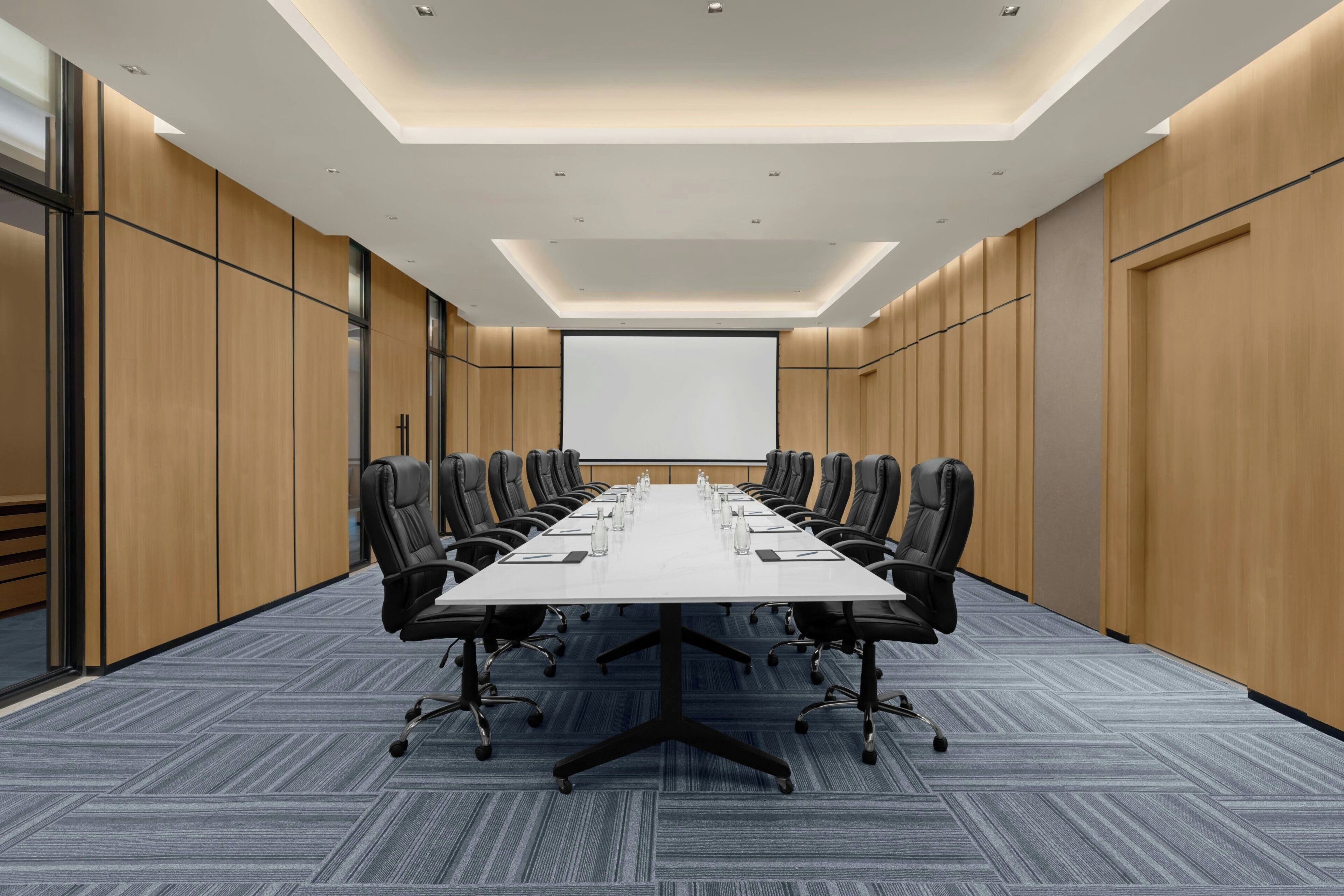A conference room with a long table, black chairs, and a projector screen.