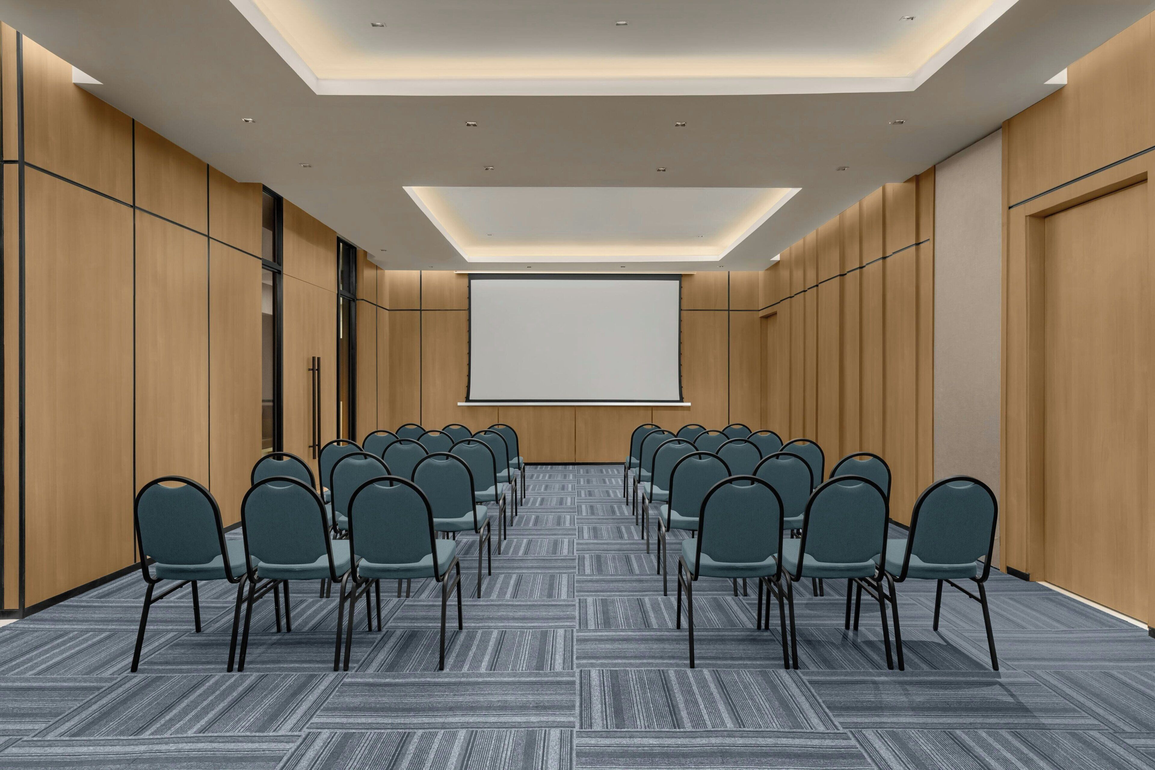 A conference room with rows of chairs facing a projection screen, wooden panel walls, and a patterned carpet.