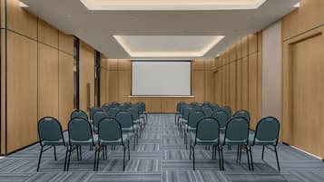 A conference room with rows of chairs facing a projection screen, wooden panel walls, and a patterned carpet.