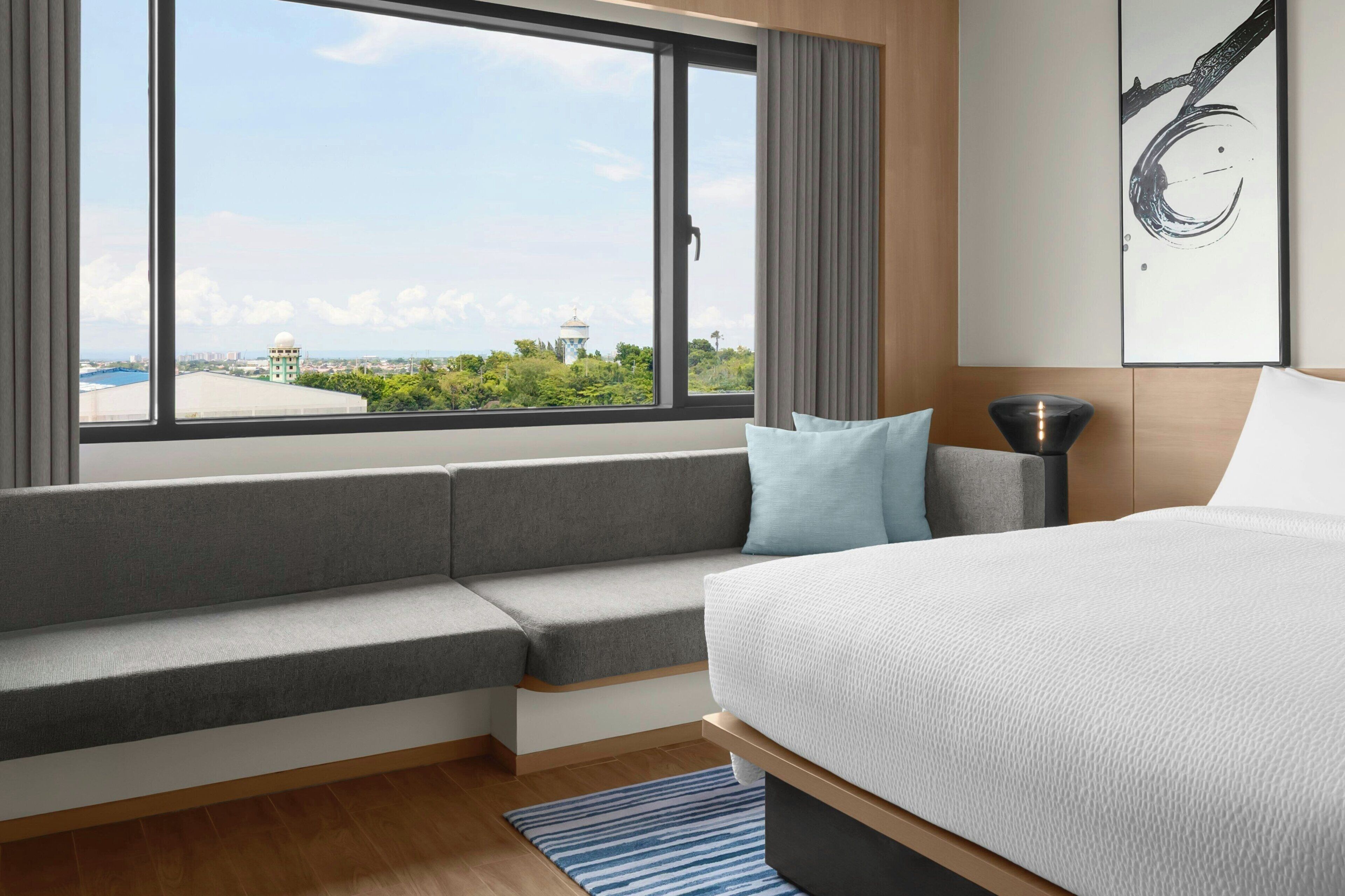 A modern hotel room with a large bed, a built-in bench, a view of the cityscape, and a painting on the wall.