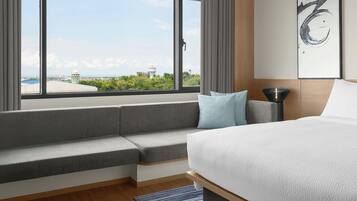 A modern hotel room with a large bed, a built-in bench, a view of the cityscape, and a painting on the wall.