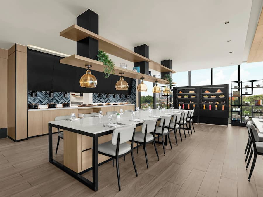 A modern kitchen with a dining area, featuring a long table set for six, wooden cabinets, and pendant lighting.