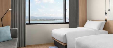 A hotel room with two beds, a desk, a chair, a window with a view of a body of water and distant landscape, and a lamp.