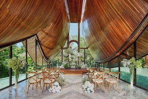 An indoor venue with a wooden ceiling, large windows, and a central stage with floral arrangements. - Hiliwatu, Bali Ubud, A Tribute Portfolio Resort (Payangan)