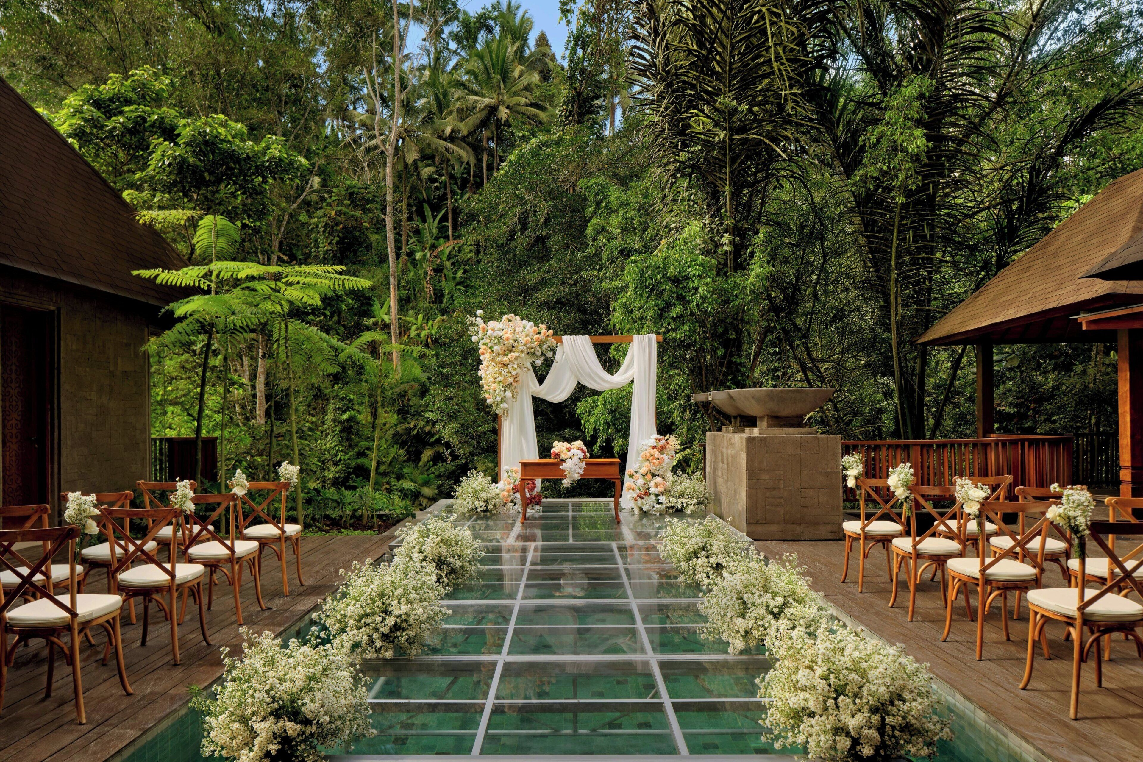 Un lieu de réception en plein air aux allures tropicales, avec une passerelle en verre menant à un autel décoré, entouré d’une végétation luxuriante et de structures en bois.