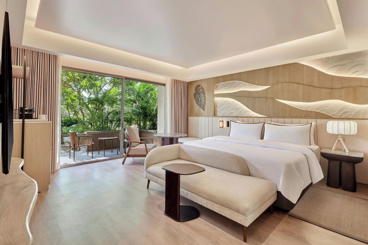 A modern hotel room with a large bed, a small round table, a sofa, a TV, and a view of greenery through the sliding glass door.