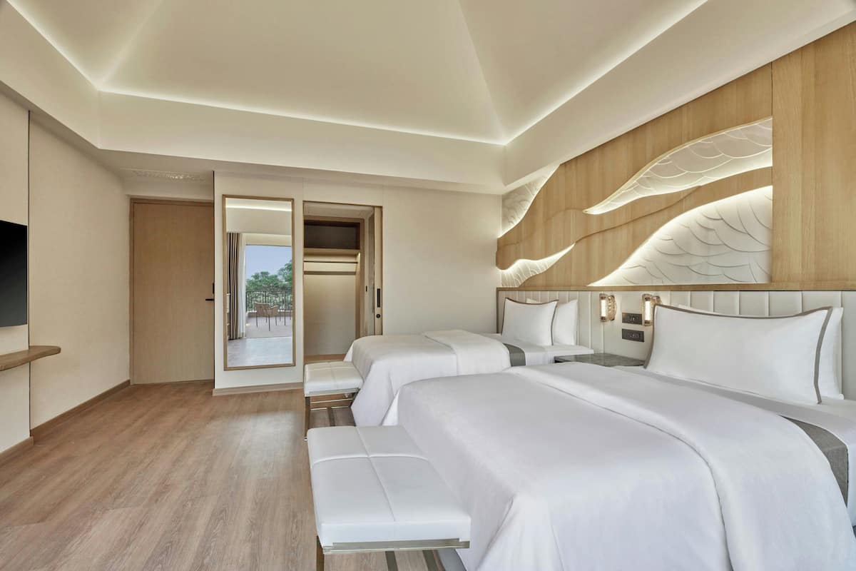 A modern hotel room with three beds, a flat-screen TV, and a large window offering a view of the outdoors.