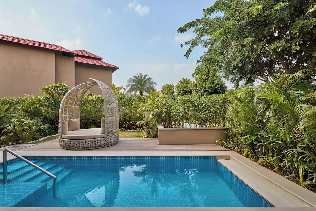 A swimming pool with a circular seating area, surrounded by lush greenery and a building with a red roof.