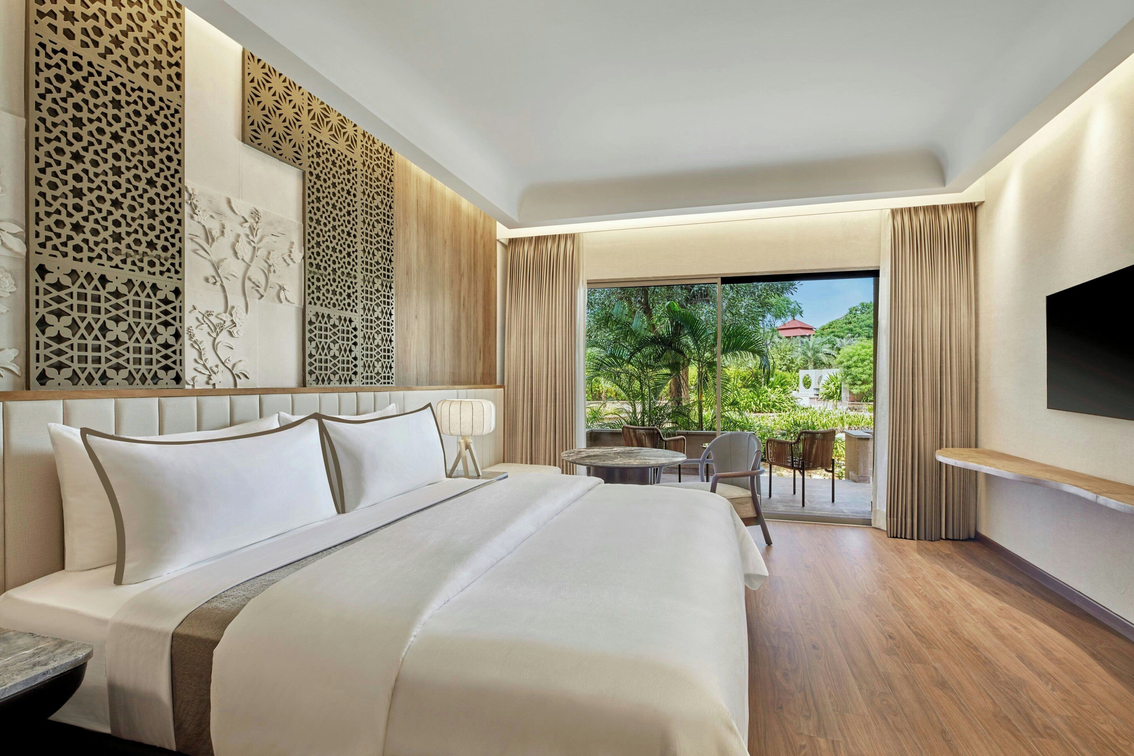 Una habitación de hotel moderna con una cama grande, un televisor montado en la pared y un balcón con vistas a zonas verdes.