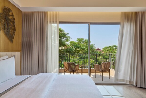 A hotel room with a large bed, a balcony with chairs, and a view of greenery.