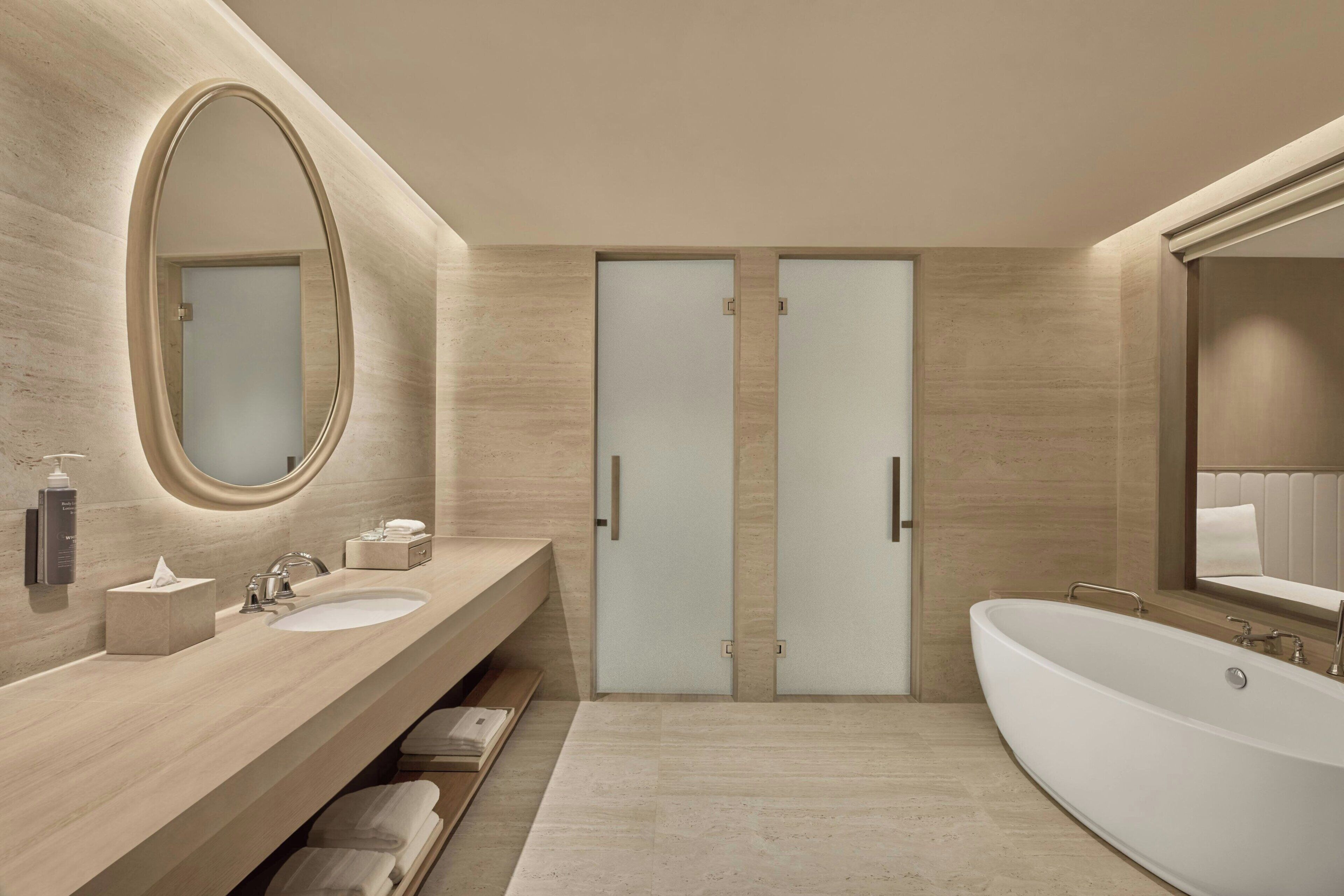 A modern bathroom with a large oval mirror, two sinks, a bathtub, and a towel rack.