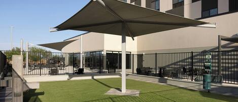 An outdoor seating area with a large umbrella, a trash bin, and a building in the background.