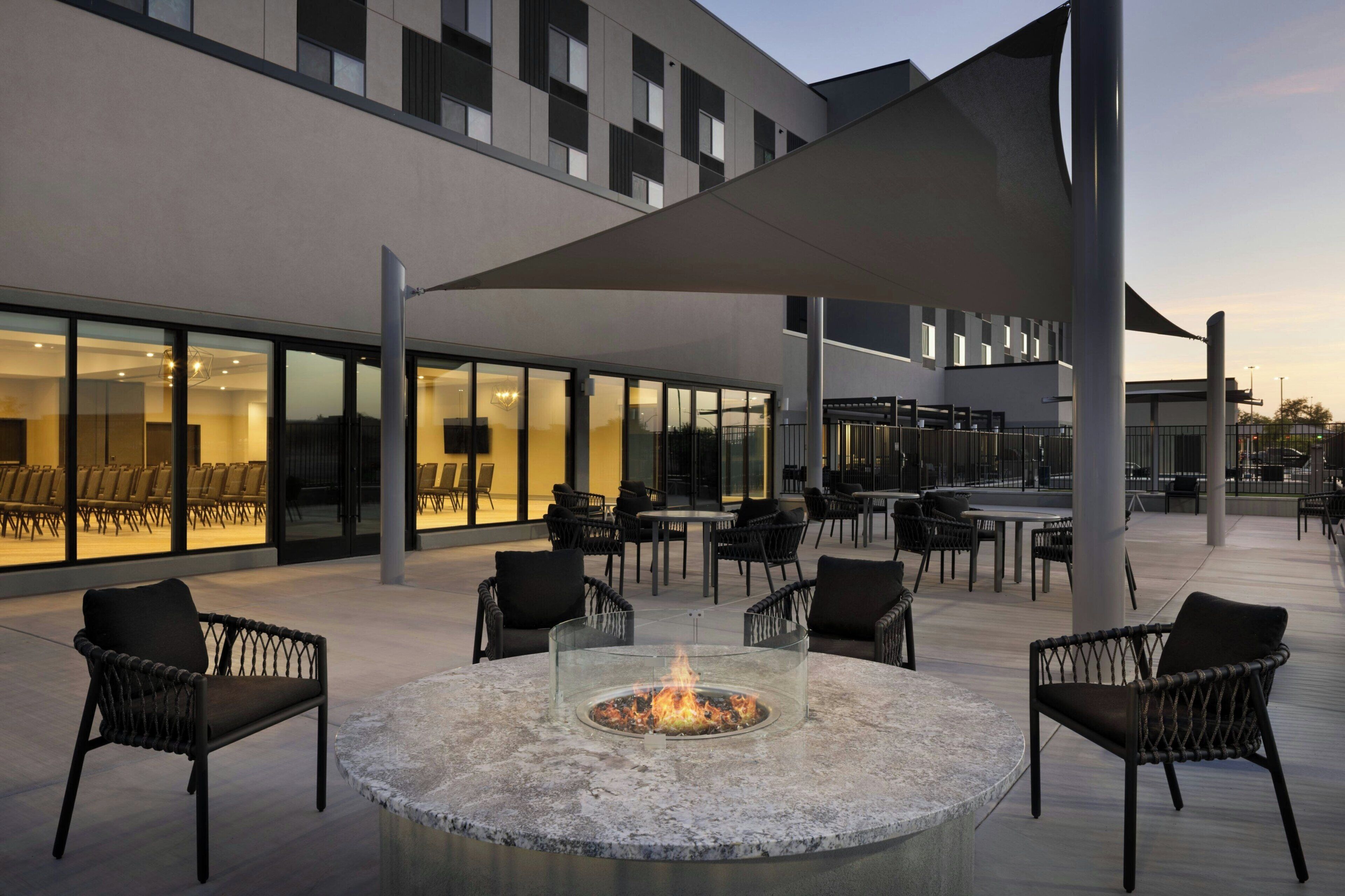 An outdoor seating area with a fire pit, chairs, and tables, adjacent to a building with large windows and a modern design.