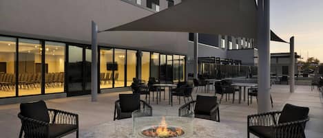 Un espace extérieur aménagé avec un foyer, des chaises et des tables, situé à côté d’un bâtiment doté de grandes fenêtres et d’un design moderne.