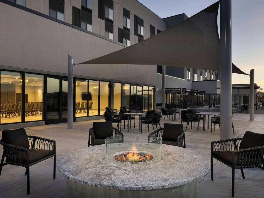 An outdoor seating area with a fire pit, chairs, and tables, adjacent to a building with large windows and a modern design.
