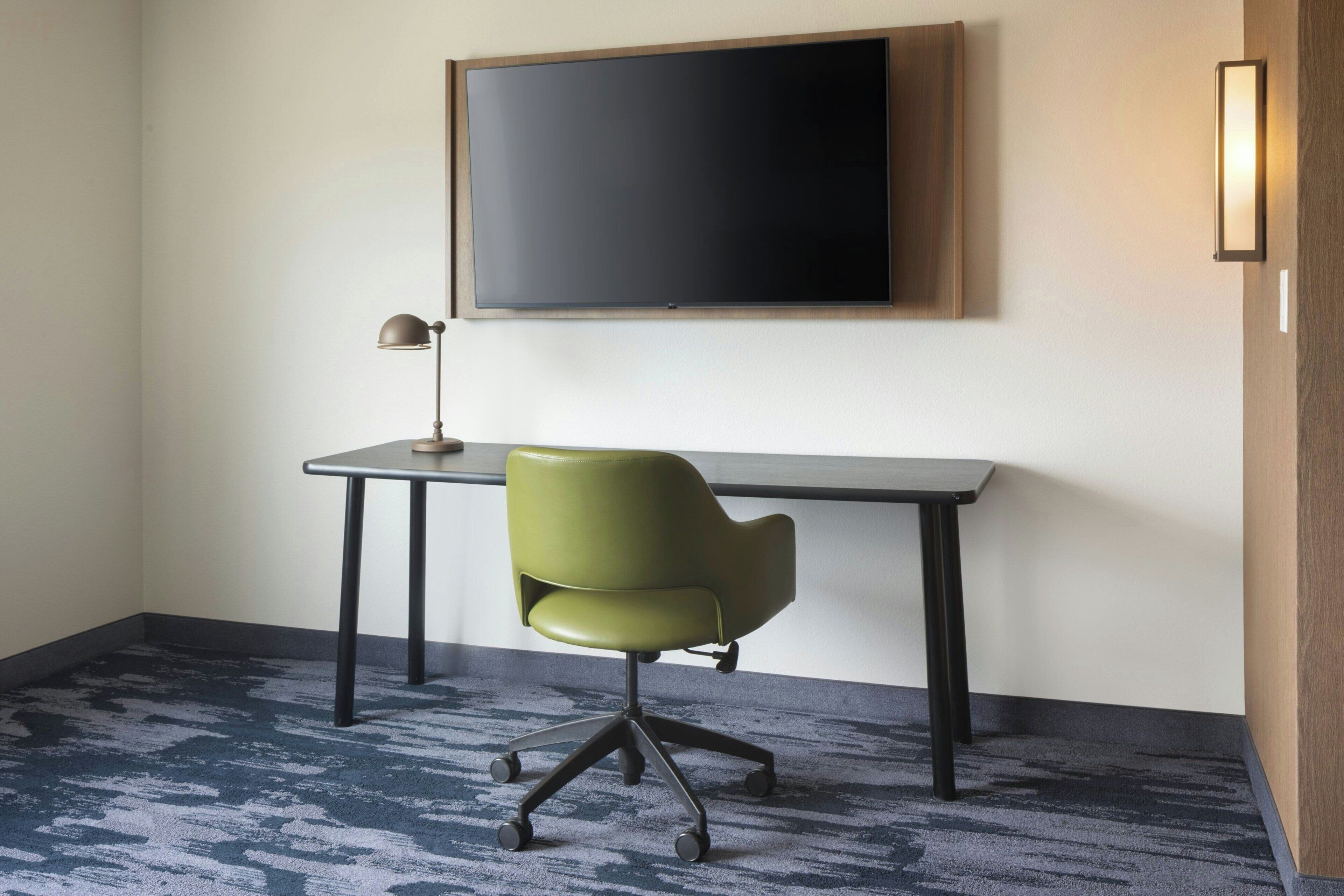 Una habitación con escritorio, silla, televisor de pantalla plana montado en la pared y piso alfombrado.