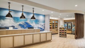 Un espace d’accueil d’hôtel avec un comptoir de réception en bois, trois lampes suspendues, une grande fresque murale sur le thème des vagues et, au fond, un présentoir réfrigéré.