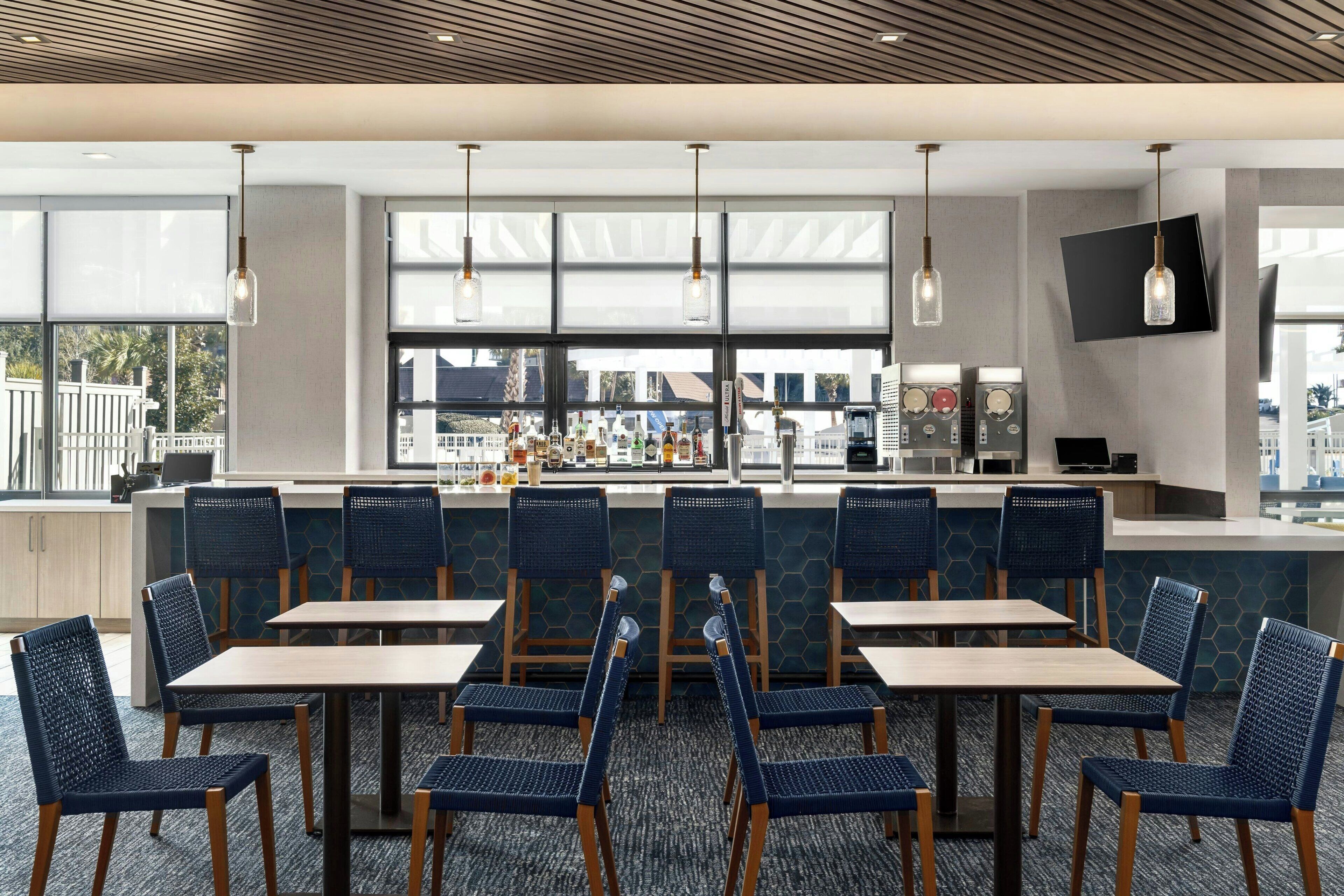 A modern dining area with tables and chairs, a bar with a counter, and large windows offering a view of the city.
