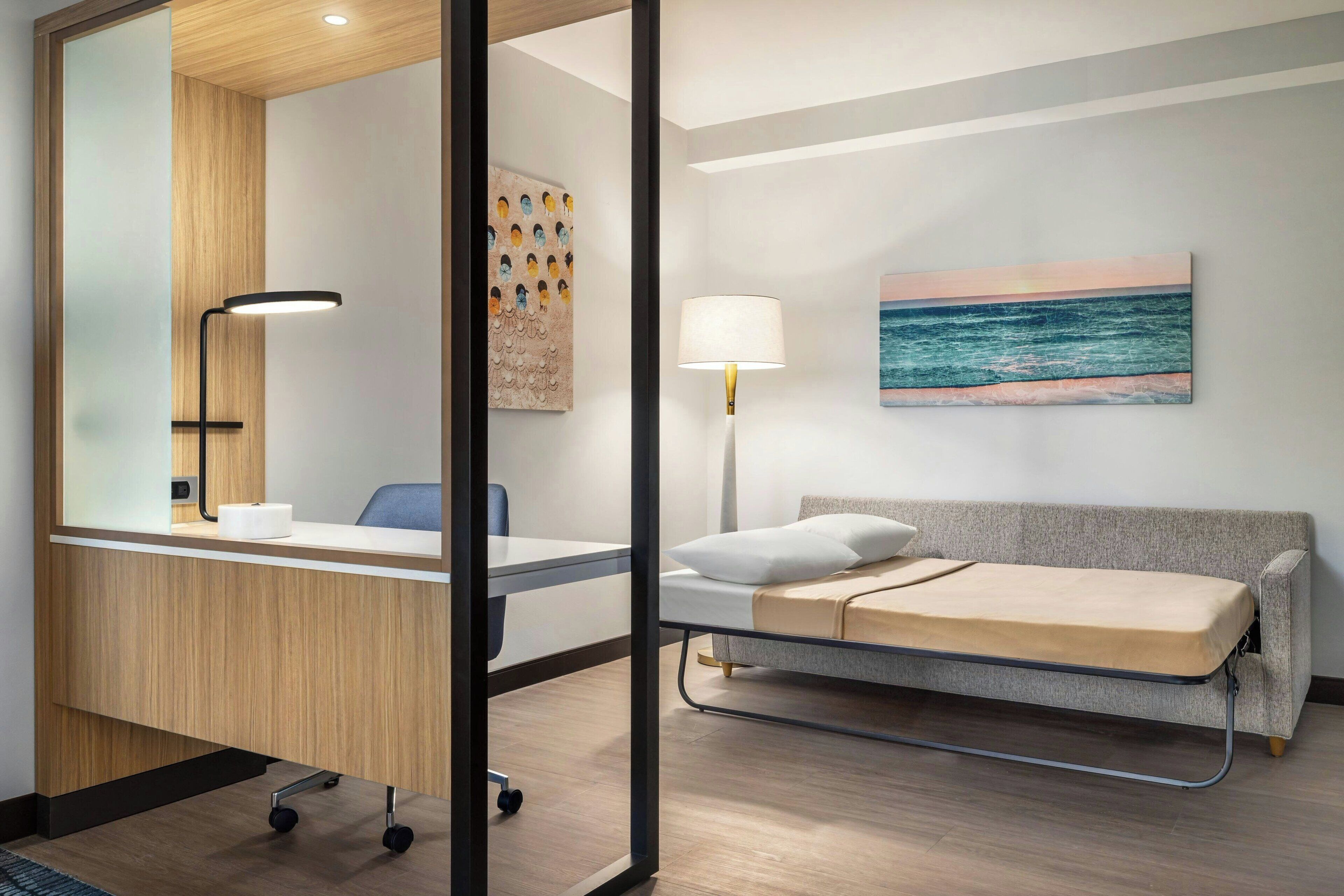 Een hotelkamer met een bureau, een bed, een lamp en een schilderij van de oceaan.
