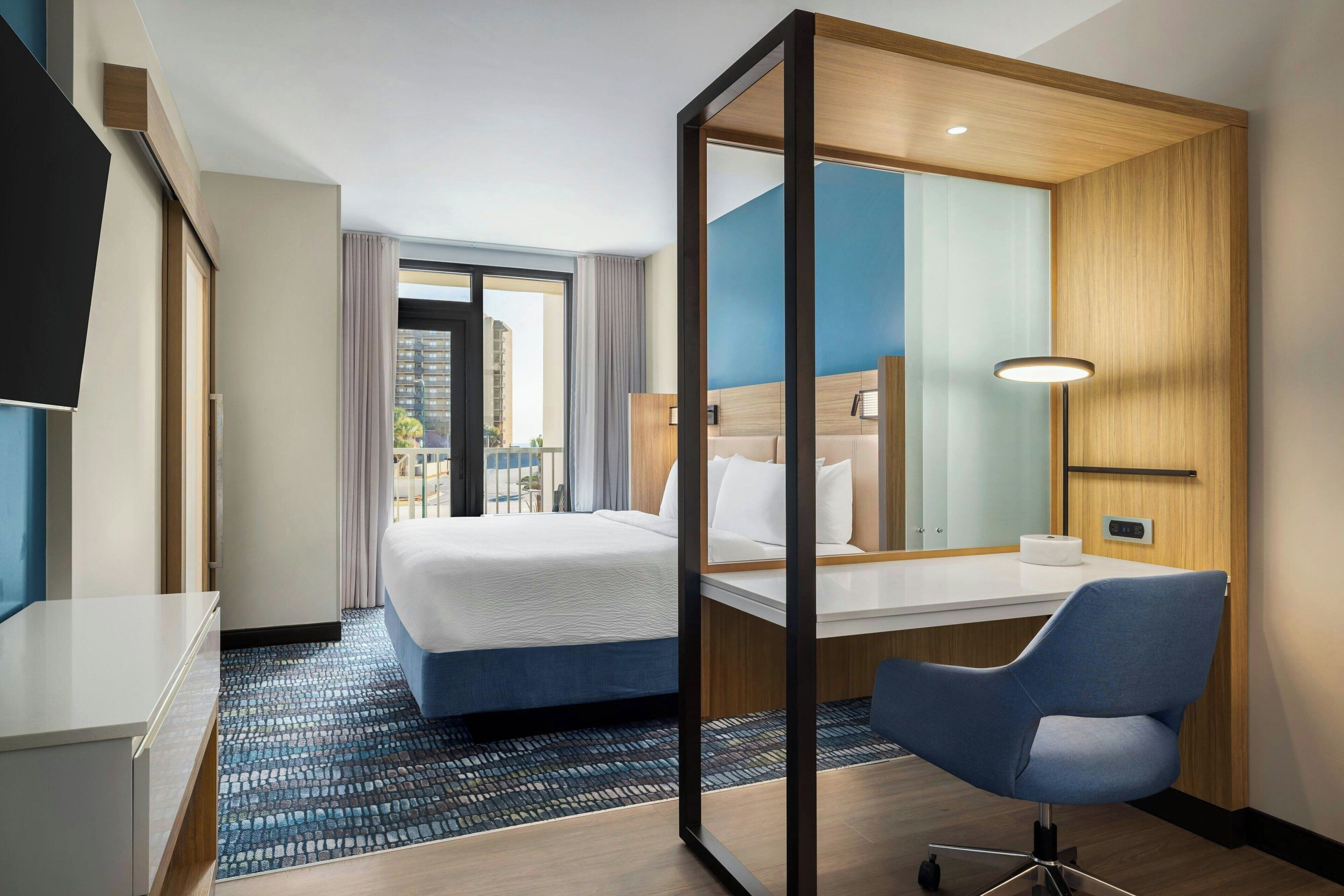 A hotel room with a bed, a desk, a chair, a TV, and a large window with a view of buildings.