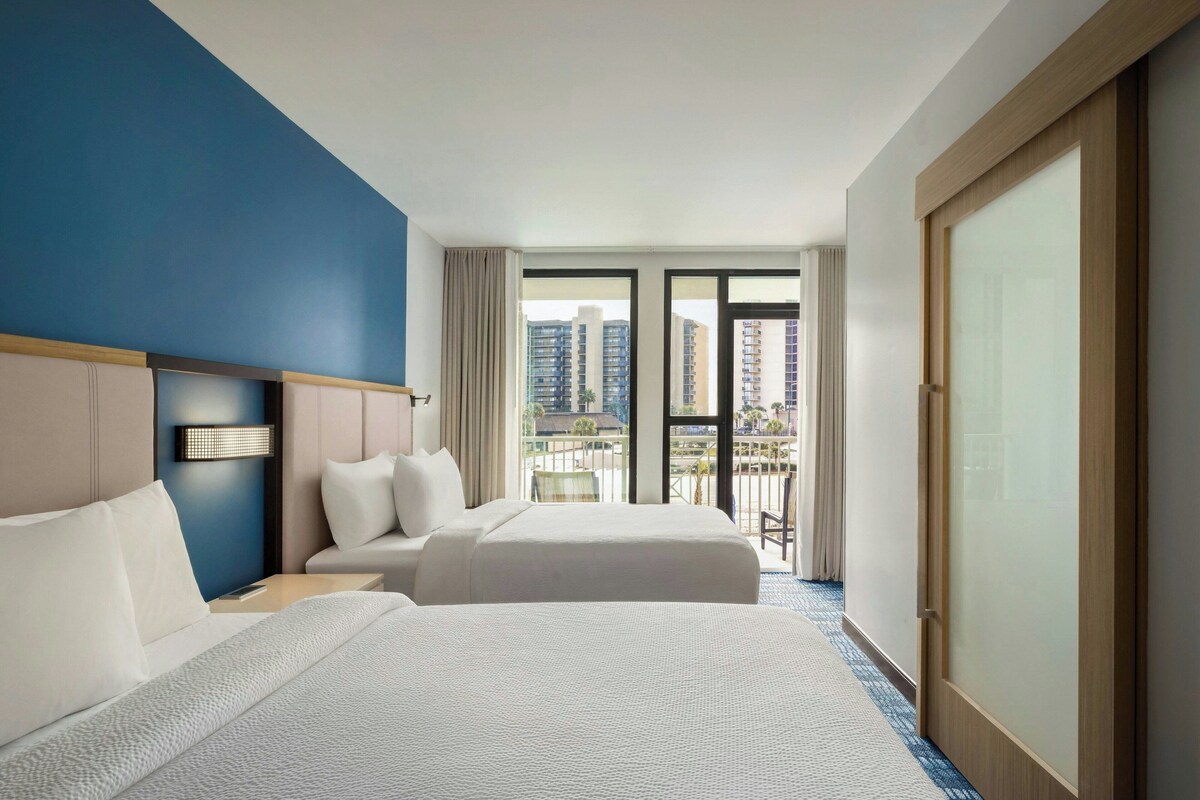 Ein Hotelzimmer mit zwei Betten, einem Nachttisch, einer Lampe und Blick auf die Stadt durch große Fenster.