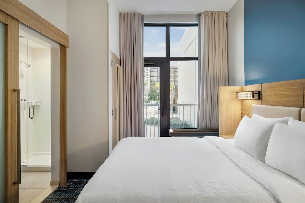 Ein Hotelzimmer mit einem großen Bett, einem Holz-Kopfteil, einem Nachttisch mit Lampe, Blick ins Freie durch eine Glastür und einer Duschkabine.