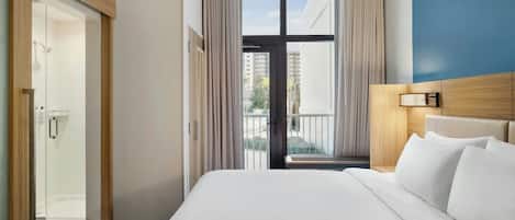 Une chambre d’hôtel avec un grand lit, une tête de lit en bois, une table de chevet avec une lampe, une vue sur l’extérieur à travers une porte vitrée et un espace douche.