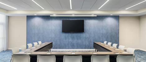 A conference room with a long table, white chairs, a flat-screen TV, and a projector on the ceiling.