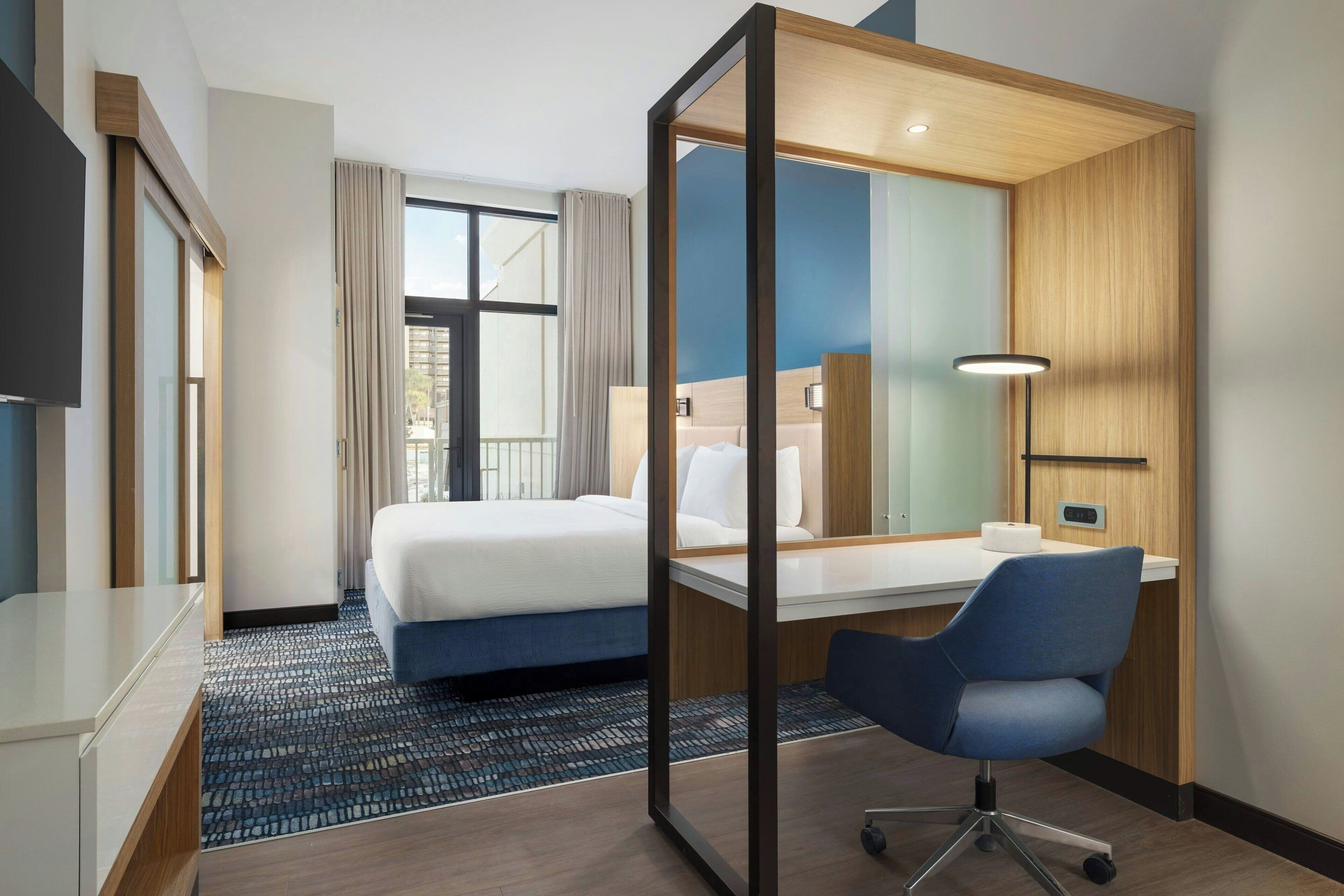 Habitación de hotel moderna con una cama grande, un escritorio y una silla. Cuenta con una ventana con cortinas, un televisor de pantalla plana y una lámpara sobre el escritorio.