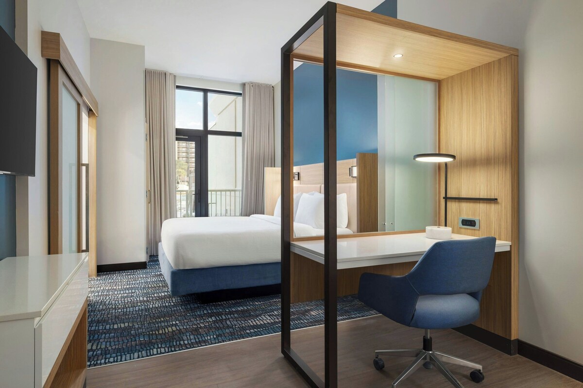 Ein modernes Hotelzimmer mit einem großen Bett, einem Schreibtisch und einem Stuhl. Ein Fenster mit Vorhängen, ein Flachbildfernseher und eine Lampe auf dem Schreibtisch sind vorhanden.