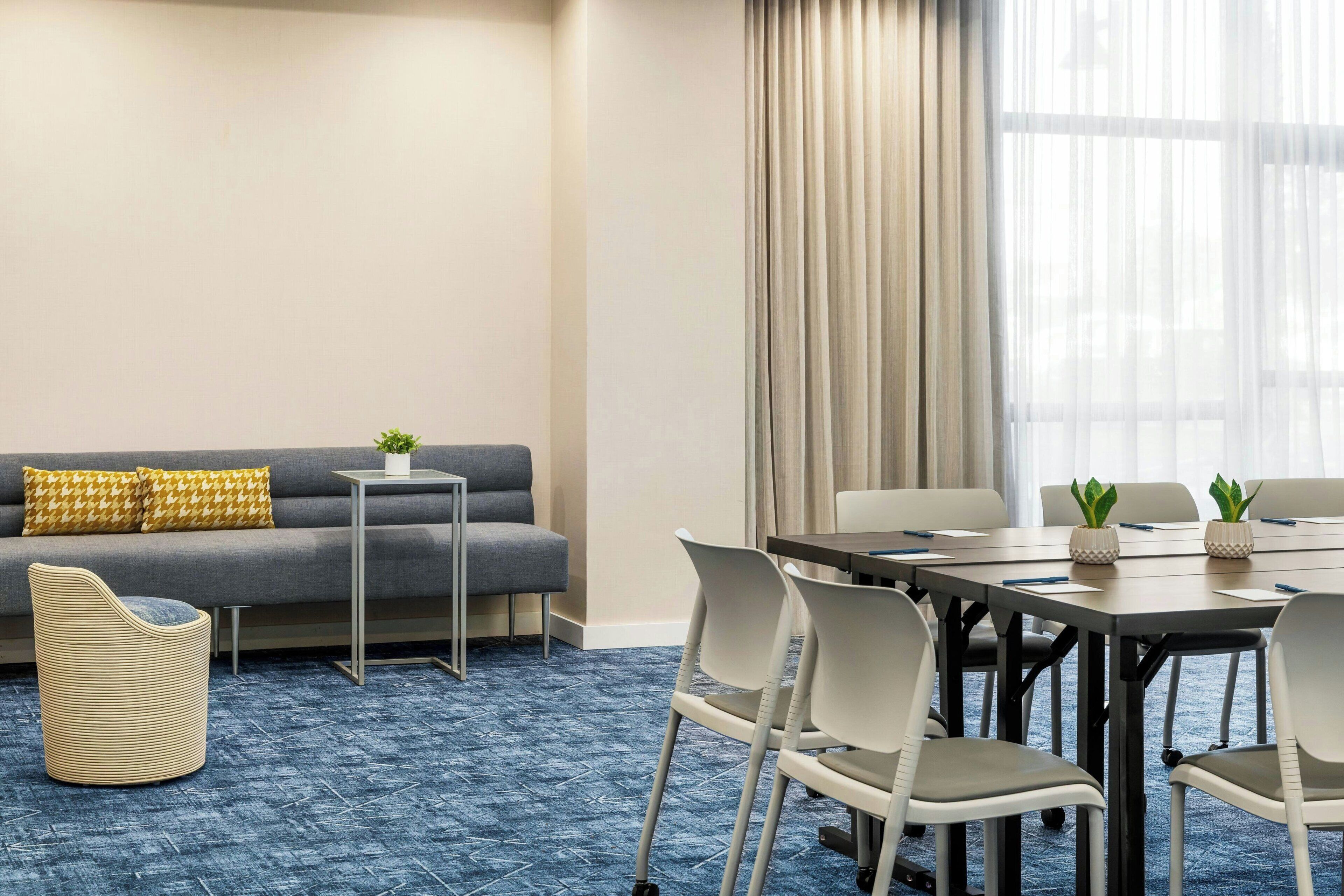 A conference room with a long table, white chairs, a sofa with yellow cushions, a small side table, and a potted plant.